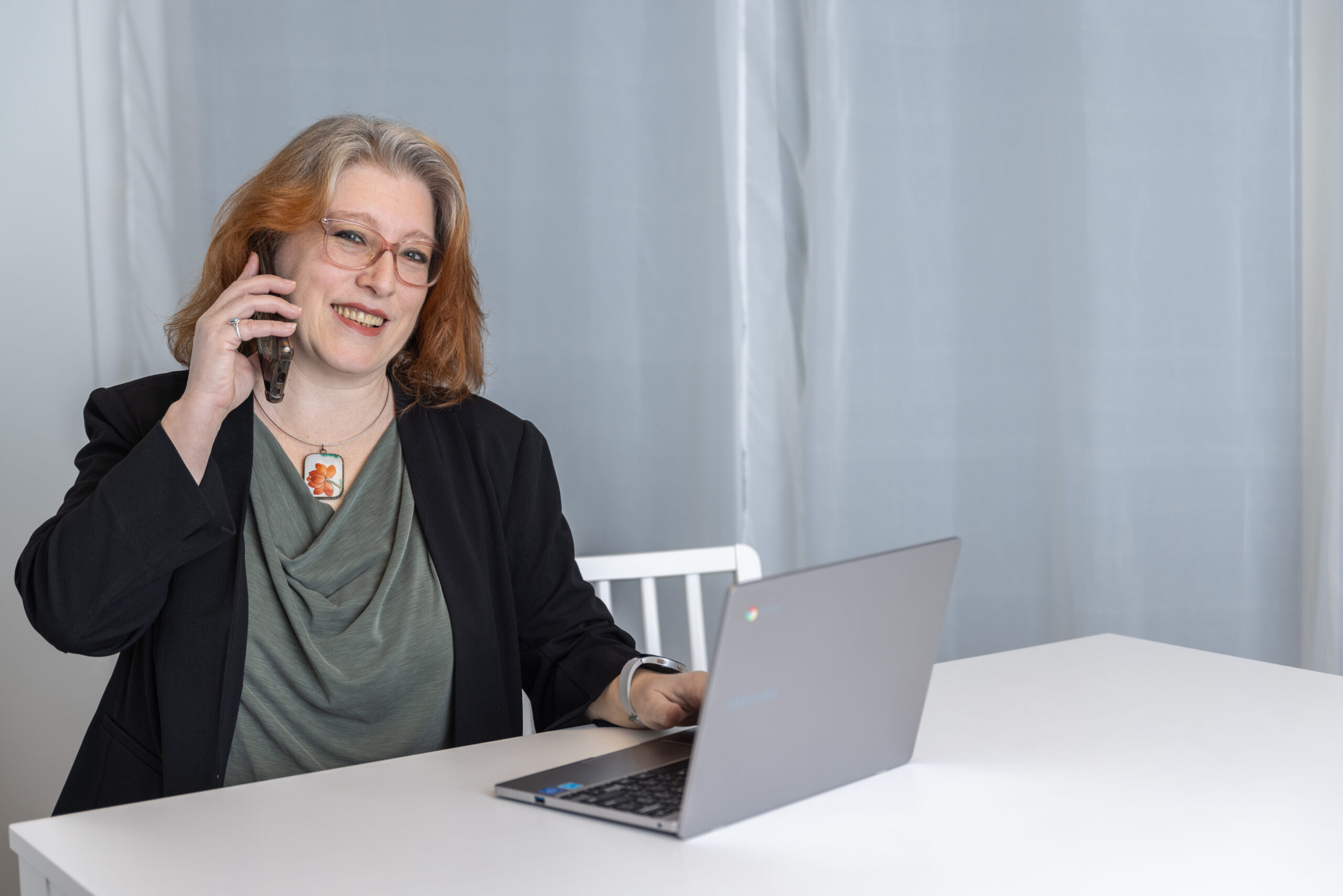 Image of Rebecca Dugas in black jacket with a cell phone at her ear and a laptop on a white desk in front of her, ready to have a Discovery Call with you!