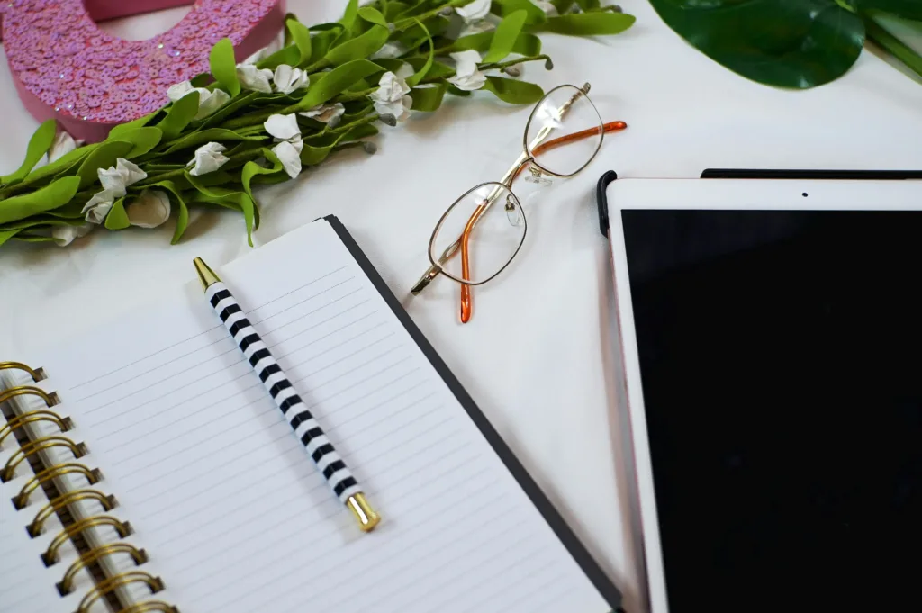 Eyeglasses, a tablet, and a sheaf of flowers sit on a white table next to a pad of paper ready to jot down newsletter topic ideas.