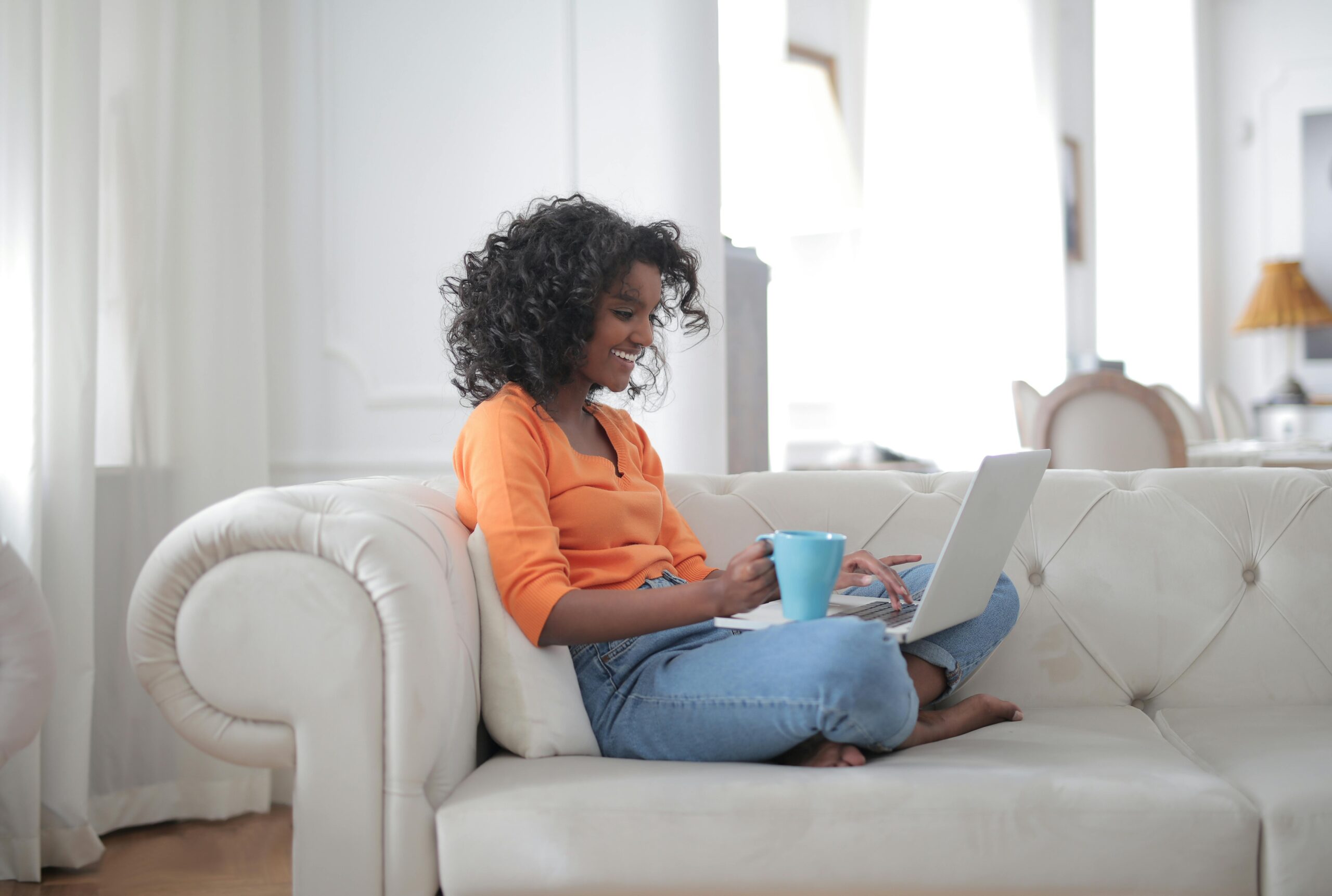 A smiling woman sitting on a white sofa with her laptop on her lap, holding a blue coffee mug, working in a bright living room. This image illustrates the ease and productivity of weekly blogging from the comfort of home.