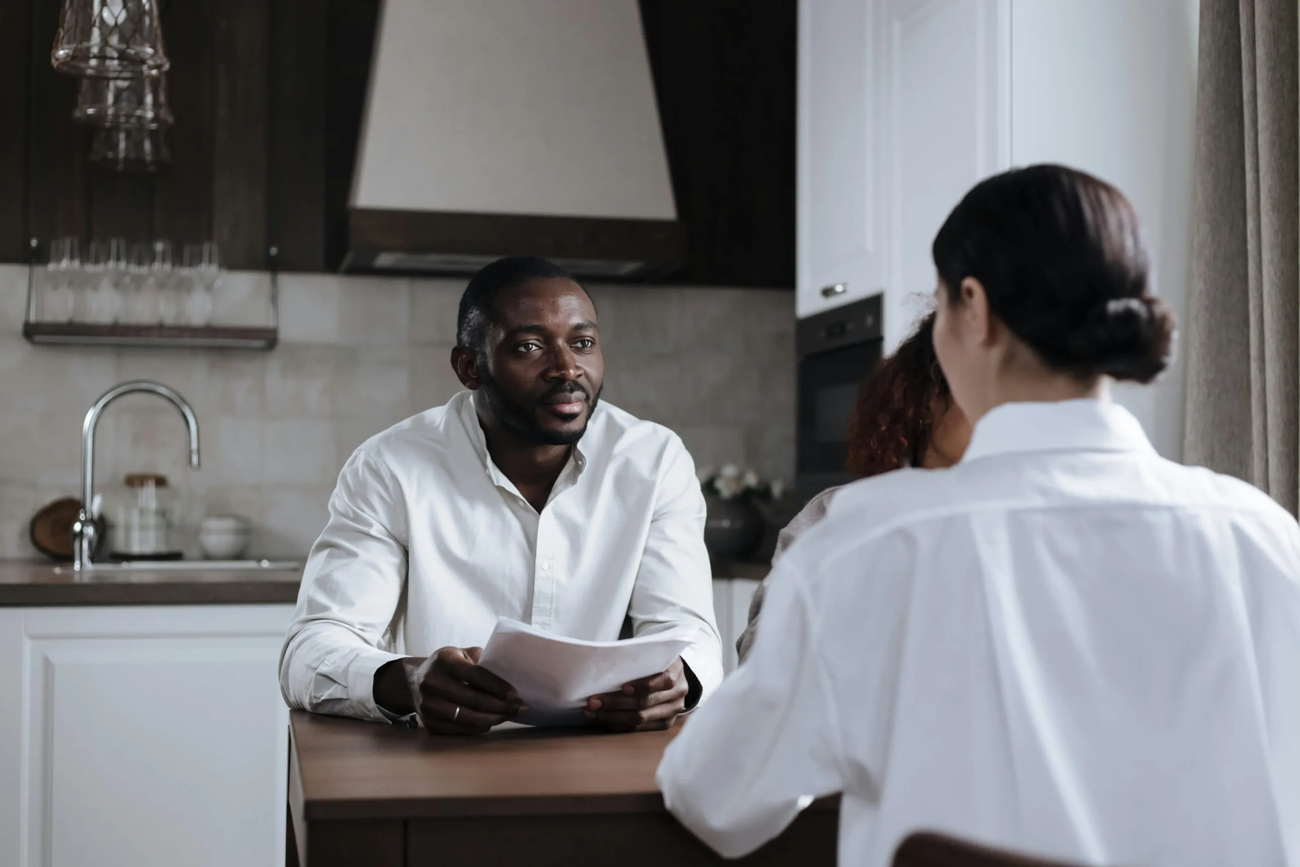 Two people having a conversation at a kitchen table; one person holds a stack of papers that could be a newsletter